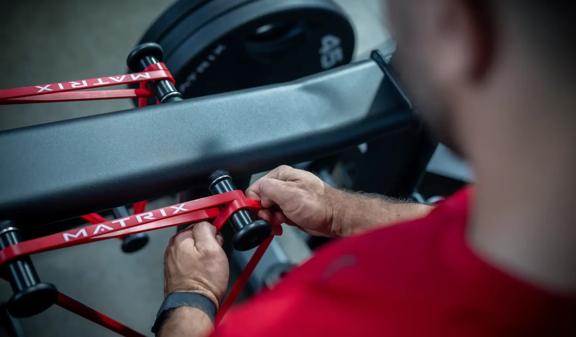 Matrix Fitness belt squat in matte black as a person adjusts a red Matrix resistance band around the machine’s rollers.